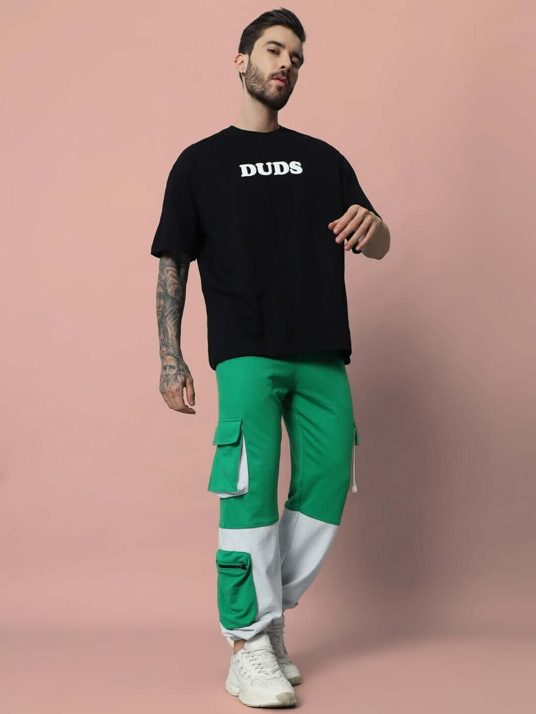 Man wearing a black oversized t-shirt with 'DUDS' print, paired with green cargo pants and white sneakers, standing in front of a pink background.
