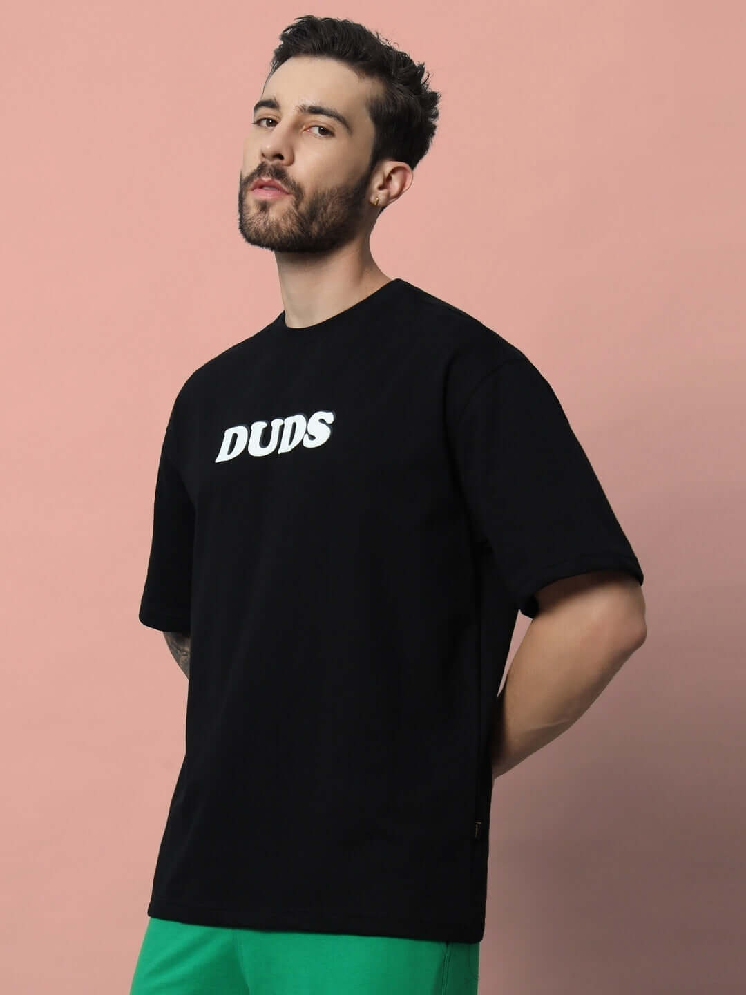 Man wearing PURE ANGEL oversized black T-shirt with white puff print, made with premium cotton fabric, styled against a pink background.