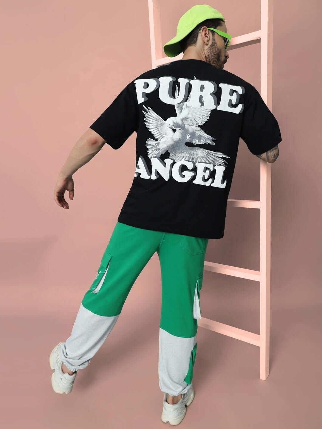 Black oversized t-shirt with 'PURE ANGEL' print, featuring a dove graphic, worn by model in green and white pants, ladder backdrop.