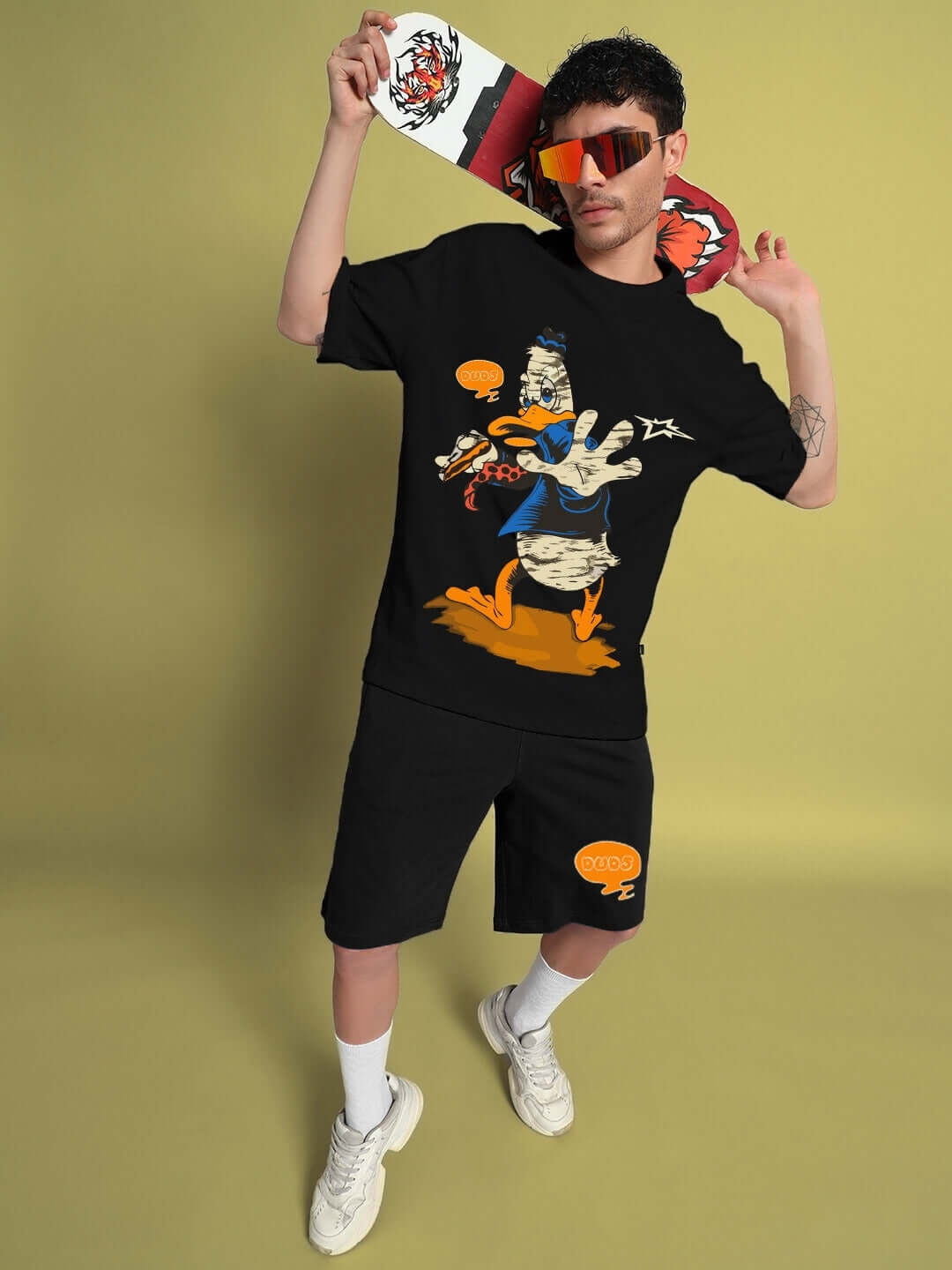 Model wearing QUACK OFF SUMMER COTTON SHORTS BLACK pairing with graphic t-shirt, showcasing streetwear style against yellow background.