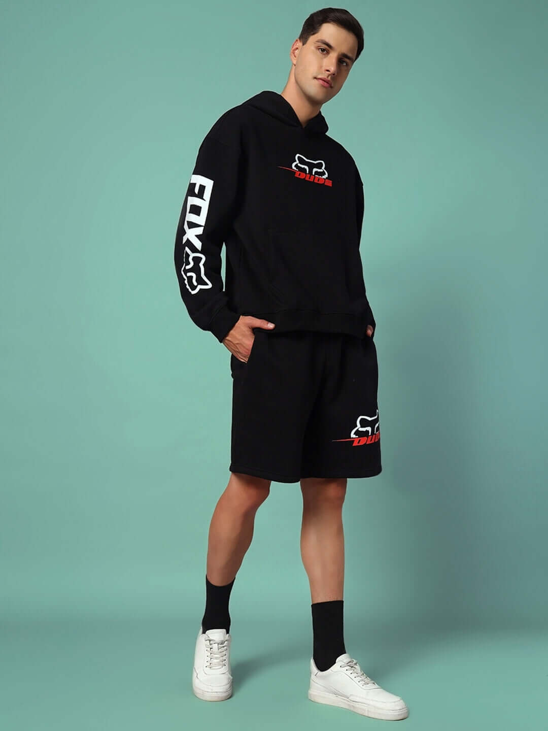 Man wearing RACING FOX FLEECE SHORTS BLACK with matching hoodie, showcasing streetwear style against a teal background.