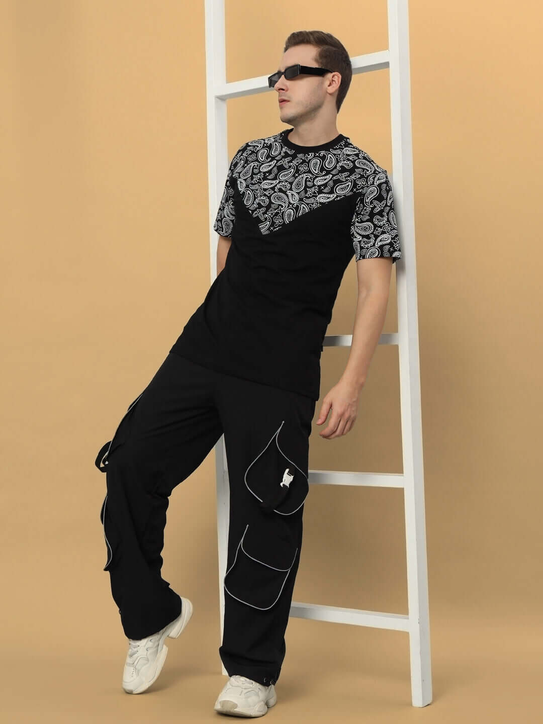 Man wearing RAGGA Reflector Parachute Cargo Pants in black, leaning on a white ladder. Casual modern style with a patterned shirt and sunglasses.