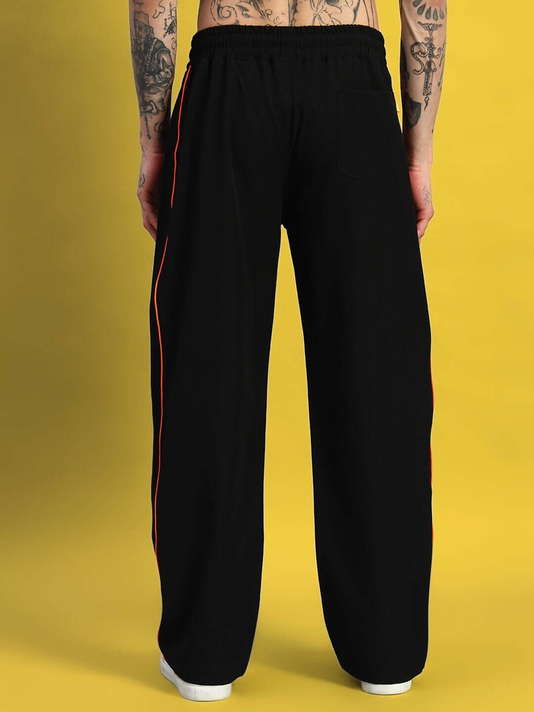 Back view of black Raider Relaxed Pant Jogger with orange side stripes, shown on a tattooed model against a yellow background.