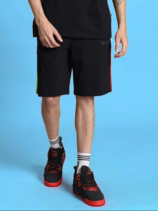 Man wearing RAINBOW REGULAR FIT SHORTS BLACK with sneakers on blue background.