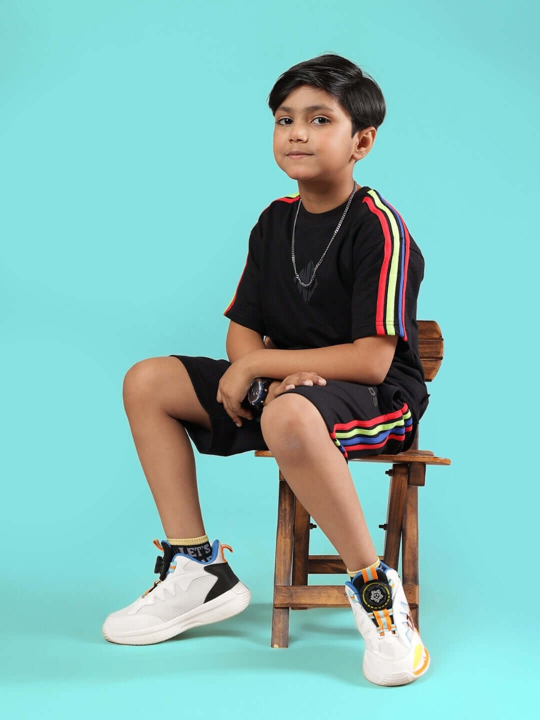 Child wearing RAINBOW SUMMER CO-ORD FOR BOYS & GIRLS BLACK sitting on a chair, showcasing rainbow-striped design, bright sneakers, and casual style.