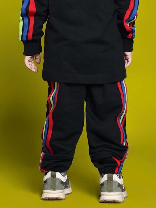 Child wearing black jogger set with rainbow stripes, showcasing back view against a yellow background.