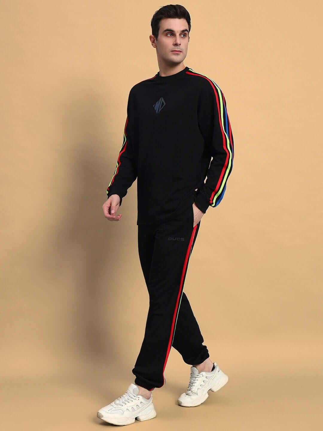 Model wearing RAINBOW OVERSIZED CO-ORD SET BLACK with colorful stripe details and white sneakers on a beige background.