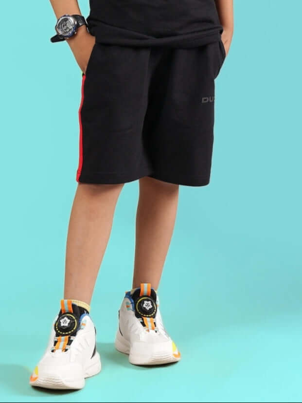 Child wearing RAINBOW SHORTS FOR BOYS & GIRLS BLACK with sneakers and a watch against a blue background.