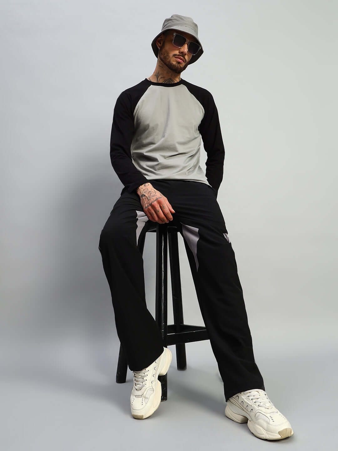 Man wearing black relaxed fit cargo pants and gray top, sitting on a stool, styled with a hat and sneakers against a gray background.