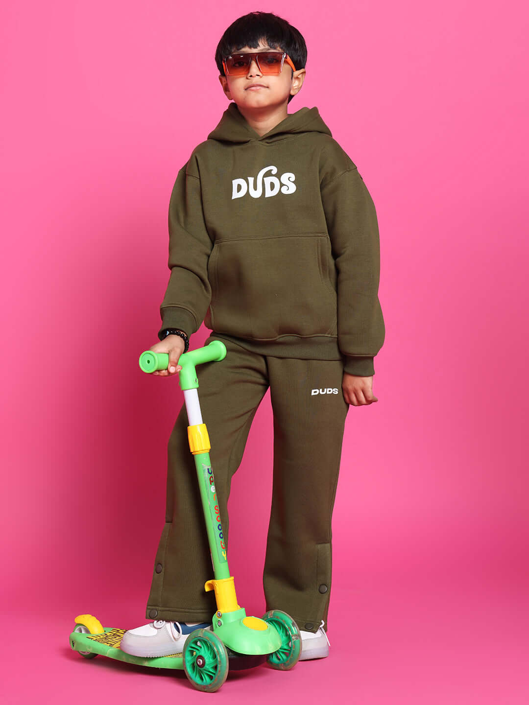 Child in olive green fleece pant and hoodie riding a green scooter against a pink background.