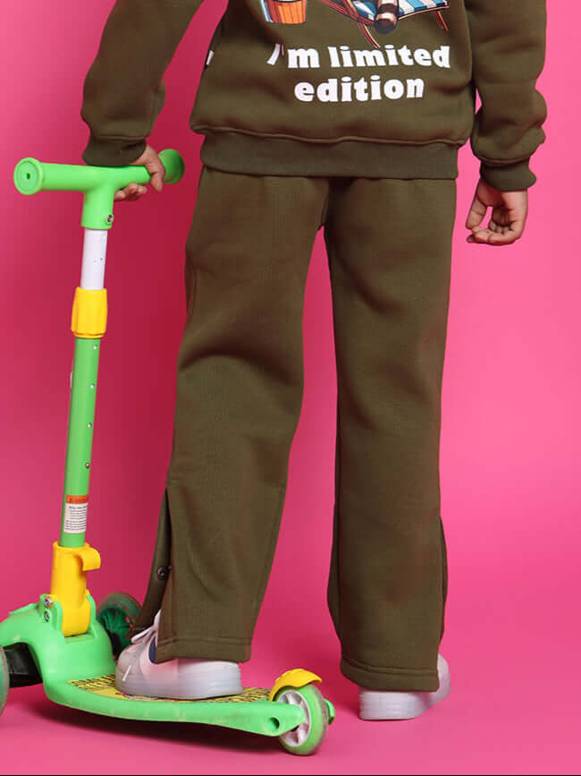 Child wearing olive green fleece pants and sweatshirt, standing next to a green scooter against a pink background.