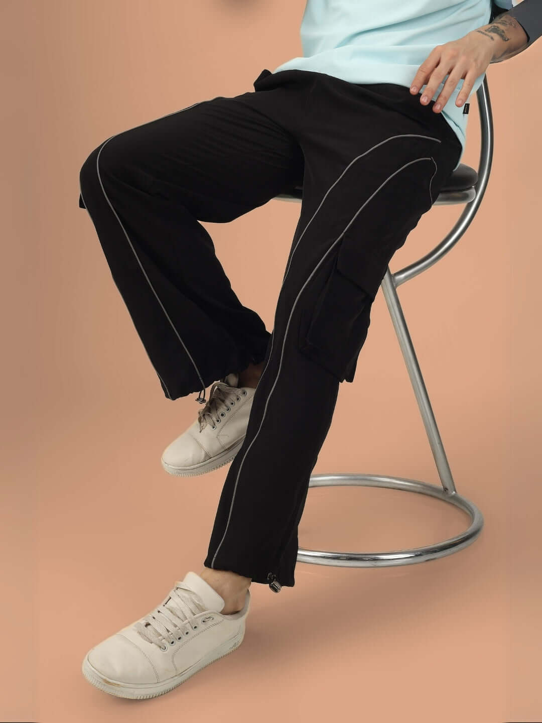 Person wearing RAY REFLECTOR Parachute Cargo Pants in black, seated on a modern chair, showcasing customizable fit and stylish design.