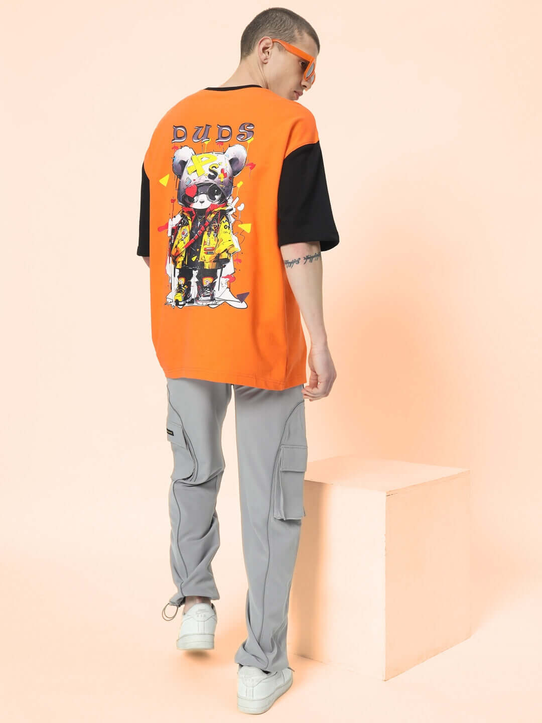 Man wearing RAY REFLECTOR parachute cargo pants in grey and an orange graphic t-shirt with a block platform backdrop.
