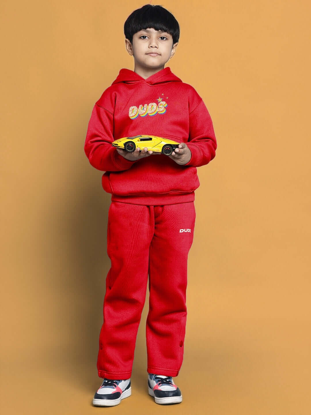 Child wearing a red Born to Shine jogger set, holding a toy car, against a yellow background.