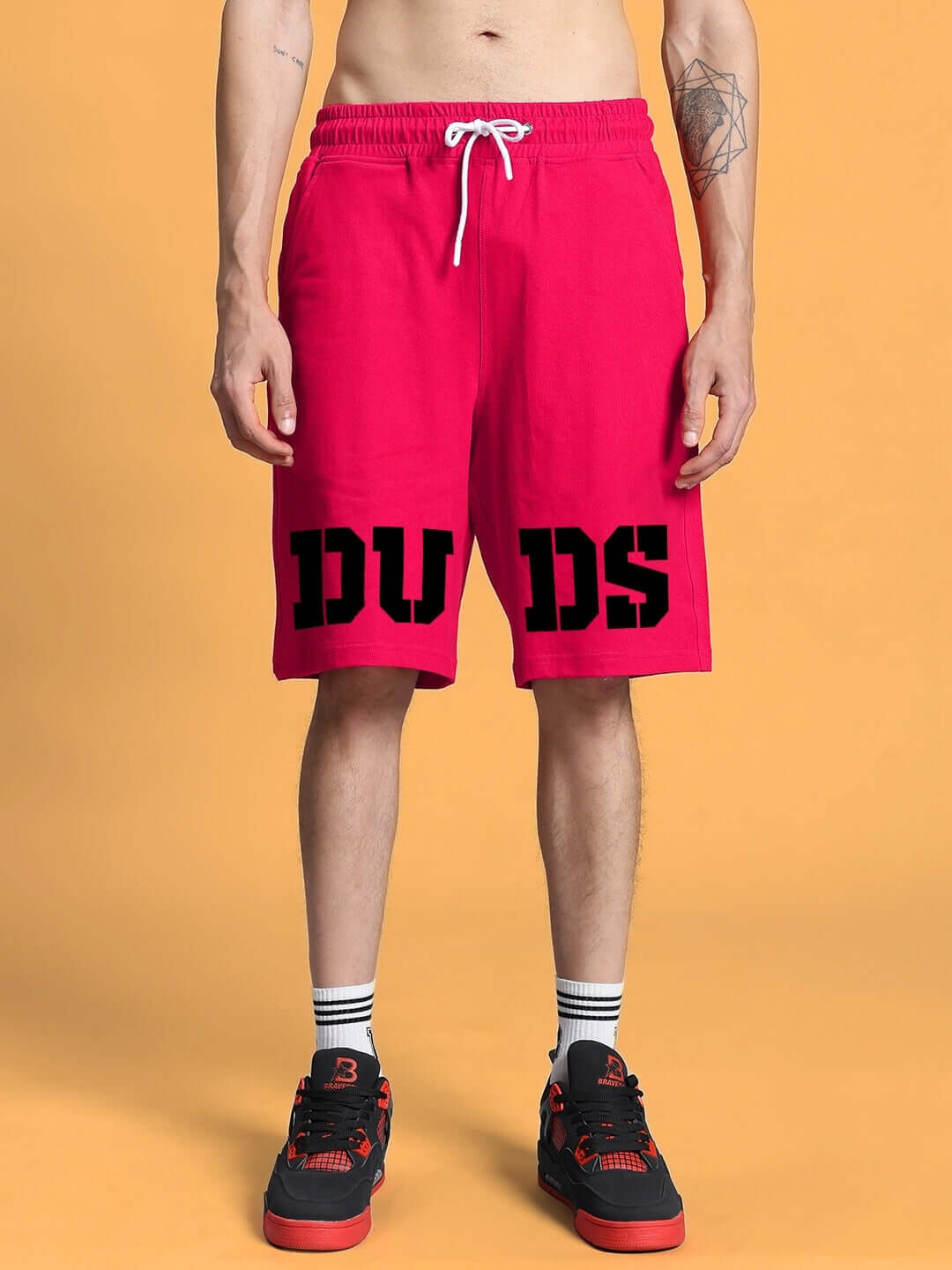 Man wearing red shorts with black "DUDS" lettering and black sneakers on orange background.