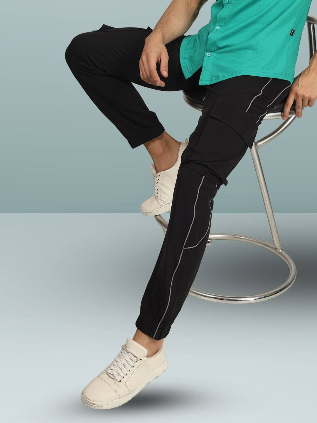 Model wearing Reflector Cargo Pants 2.0 in black, shown seated with white sneakers, highlighting custom fit option and modern design.
