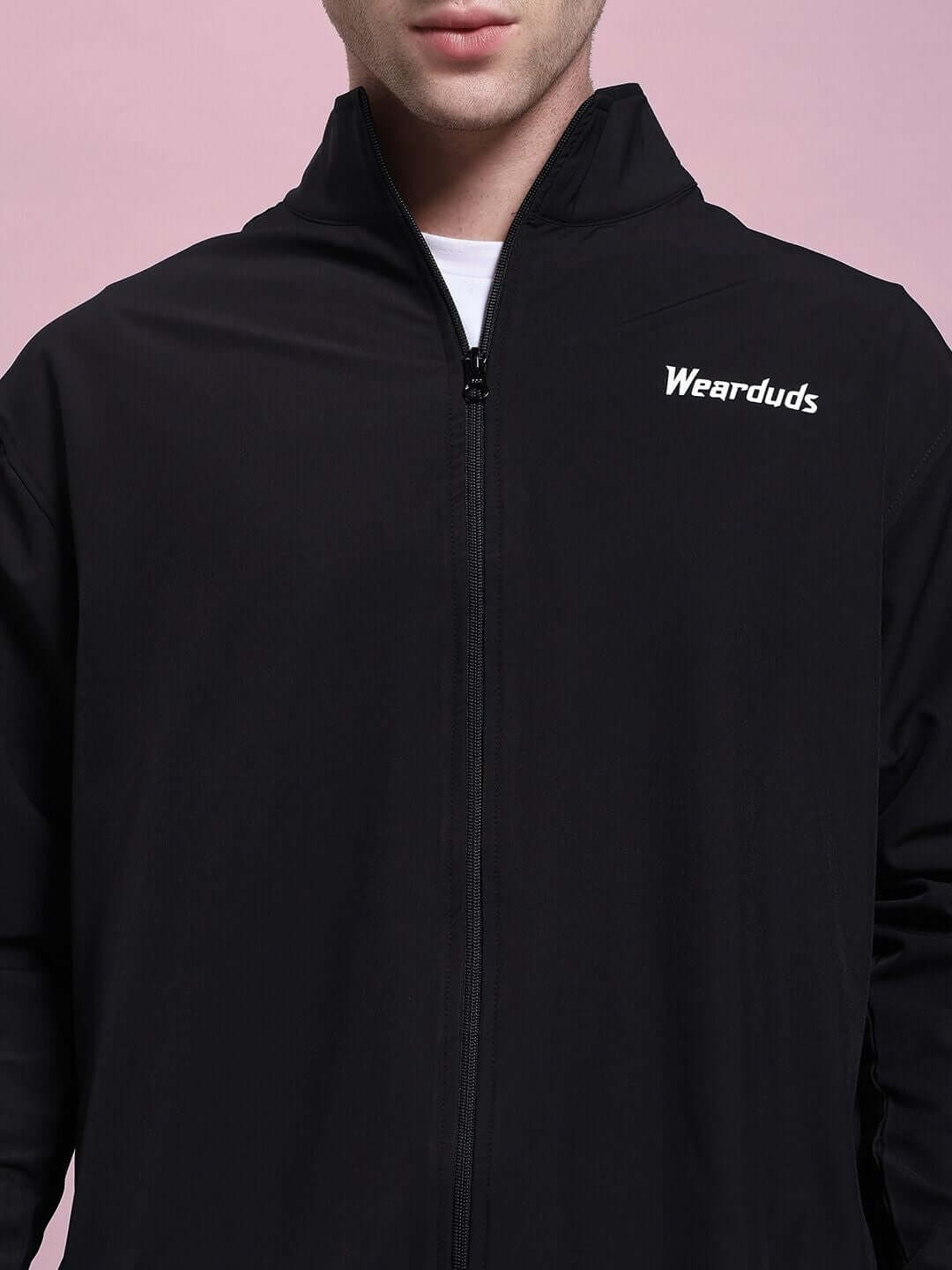 Black zipped jacket with "Wearduds" logo on chest, worn by a model, against a pink background.