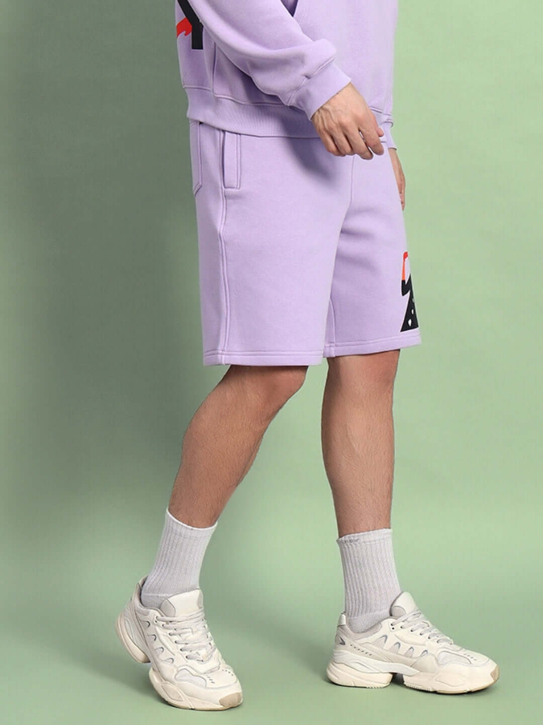 Man wearing WEARDUDS RITZY FLEECE SHORTS LILAC with white shoes on a green background.