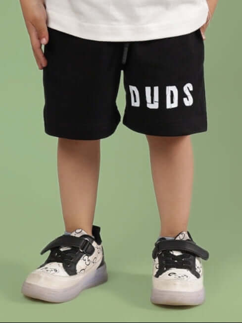 Child wearing ROBIN SHORTS FOR BOYS & GIRLS BLACK from Duds, showcasing kid's stylish shorts with shoes on a green background.