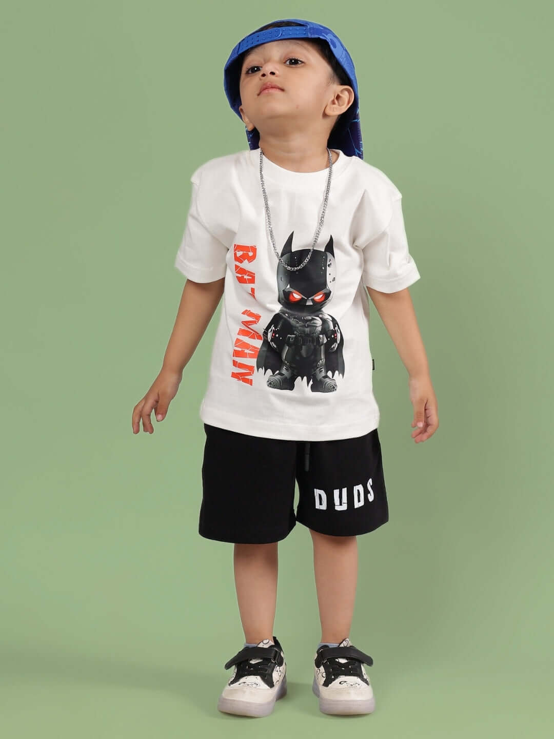 Child wearing ROBIN CO-ORD FOR BOYS & GIRLS OFF WHITE-BLACK outfit with hat, featuring cartoon and 'DUDS' printed shorts, against green background.
