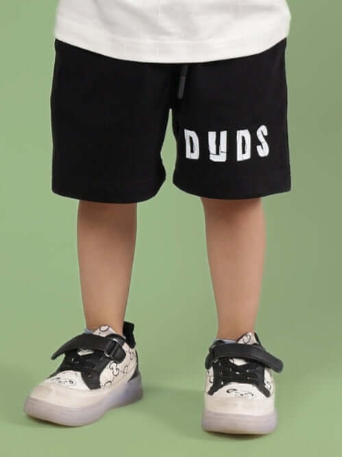 Child wearing ROBIN SHORTS FOR BOYS & GIRLS BLACK from DUDS, standing against a green background.