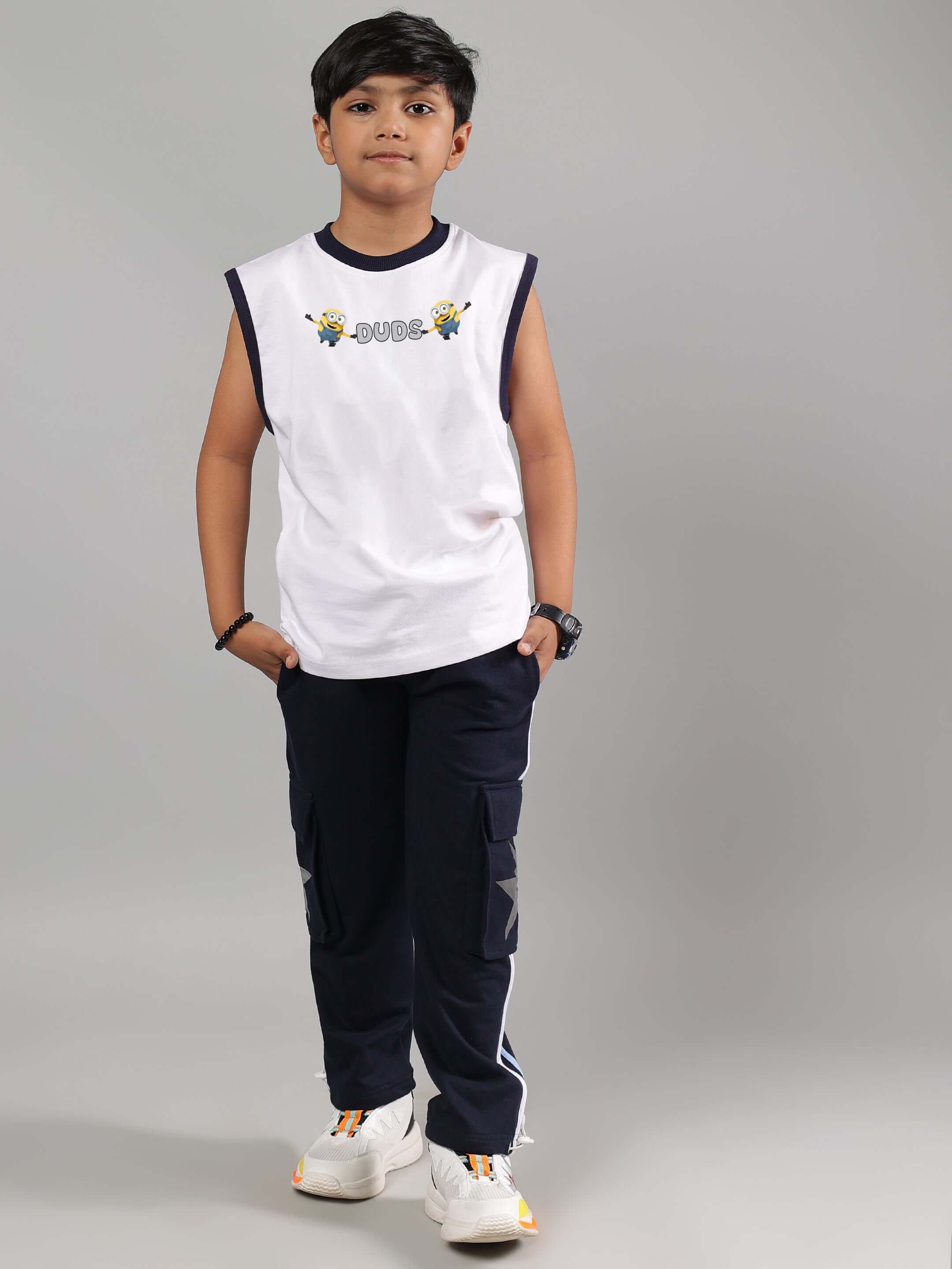 Boy wearing a white sleeveless Robo Minions T-shirt from DUDS, paired with black cargo pants and sneakers, posing against a gray background.