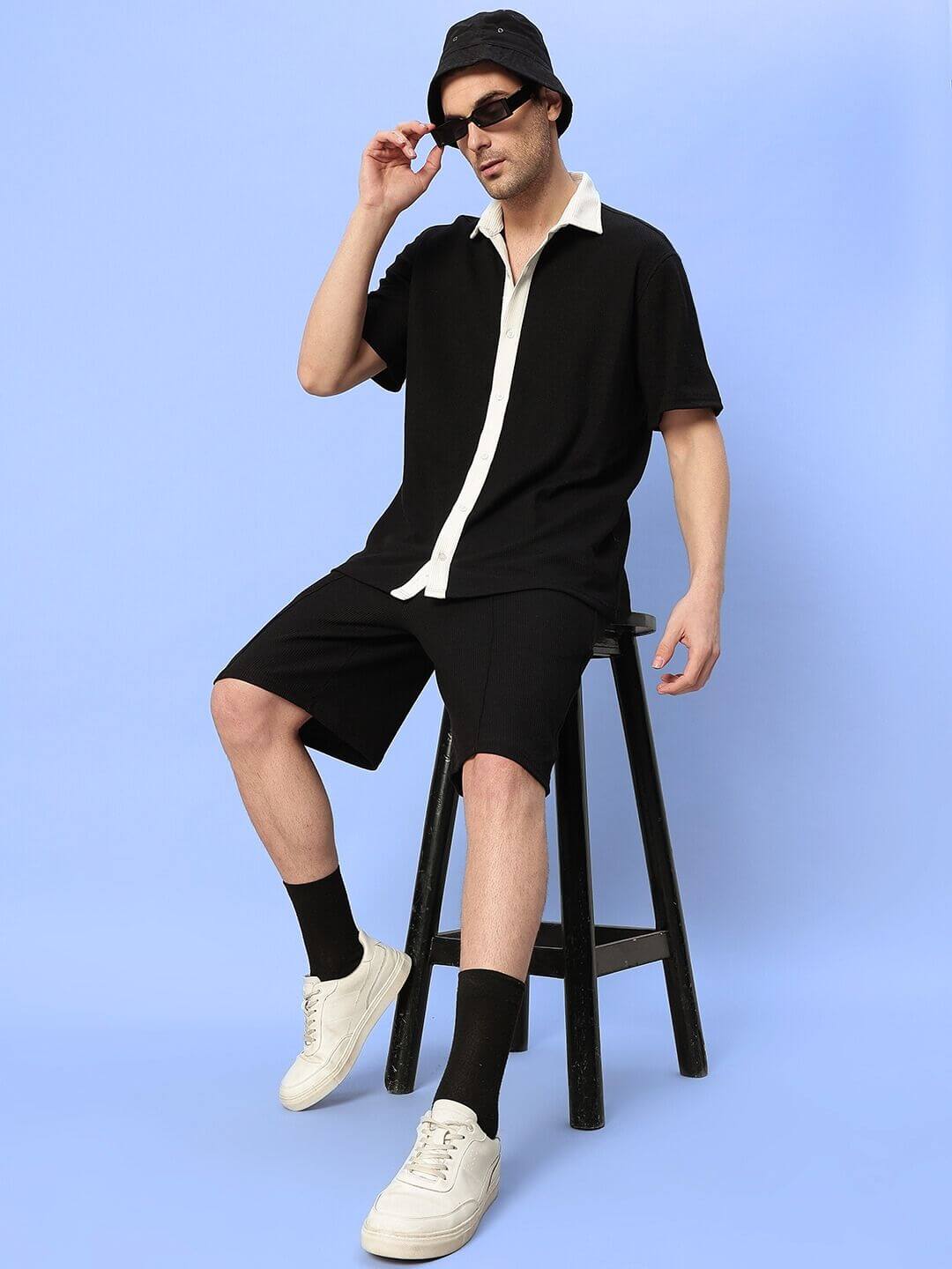 Man wearing ROBSON REGULAR FIT SHORTS BLACK with matching black shirt and bucket hat sitting on a stool against a blue background.