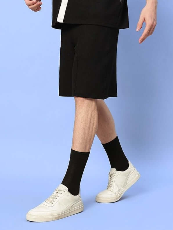 Person wearing ROBSON REGULAR FIT SHORTS BLACK, showcasing casual style with white sneakers and black socks on a blue background.
