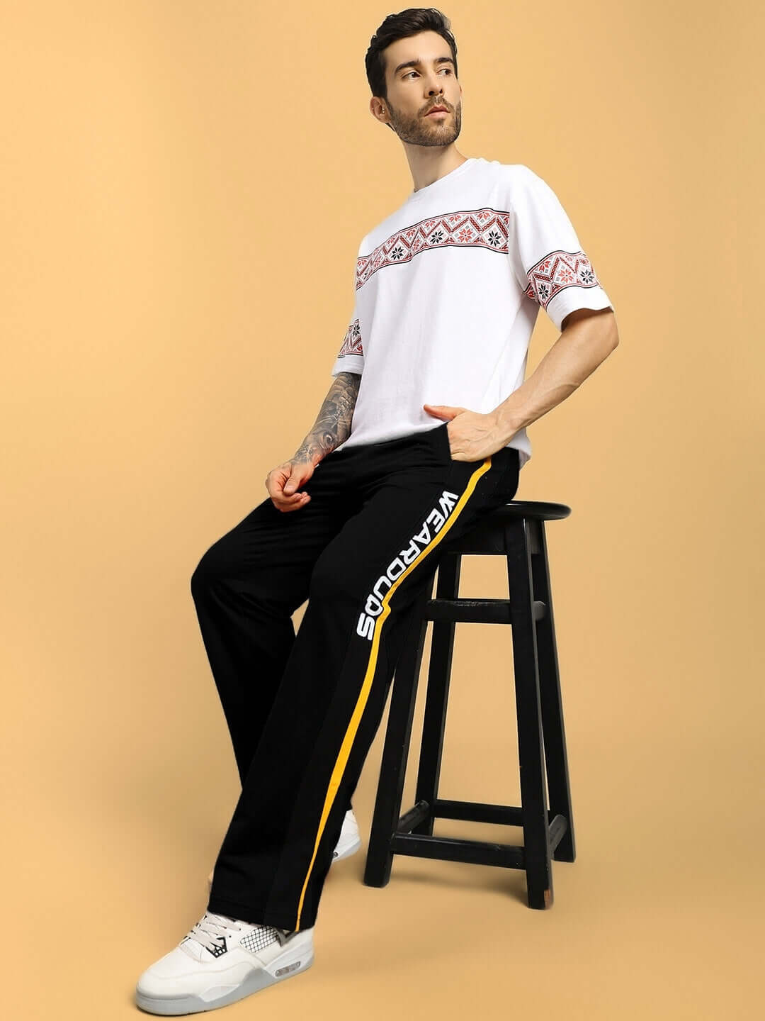 Man wearing ROGER RELAXED-FIT JOGGERS BLACK with a patterned white t-shirt, seated on a stool, showing casual streetwear style.