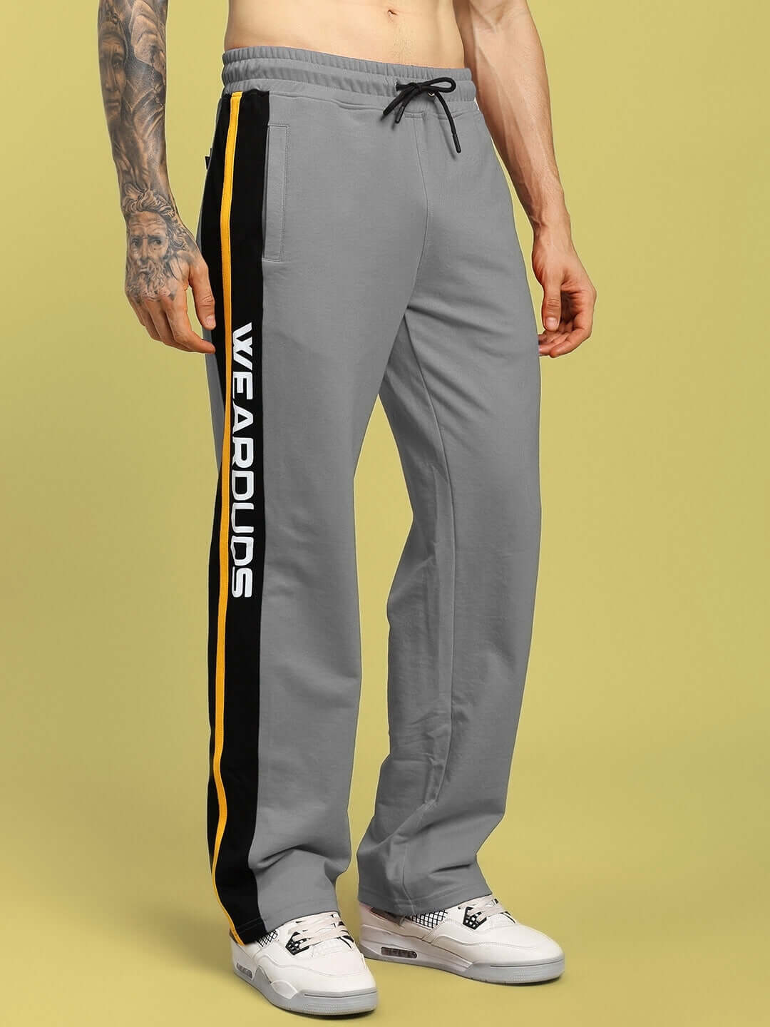 Alt Tag: Grey ROGER relaxed-fit joggers with side stripe design, crafted by DUDS, displayed on a model standing against a yellow background.