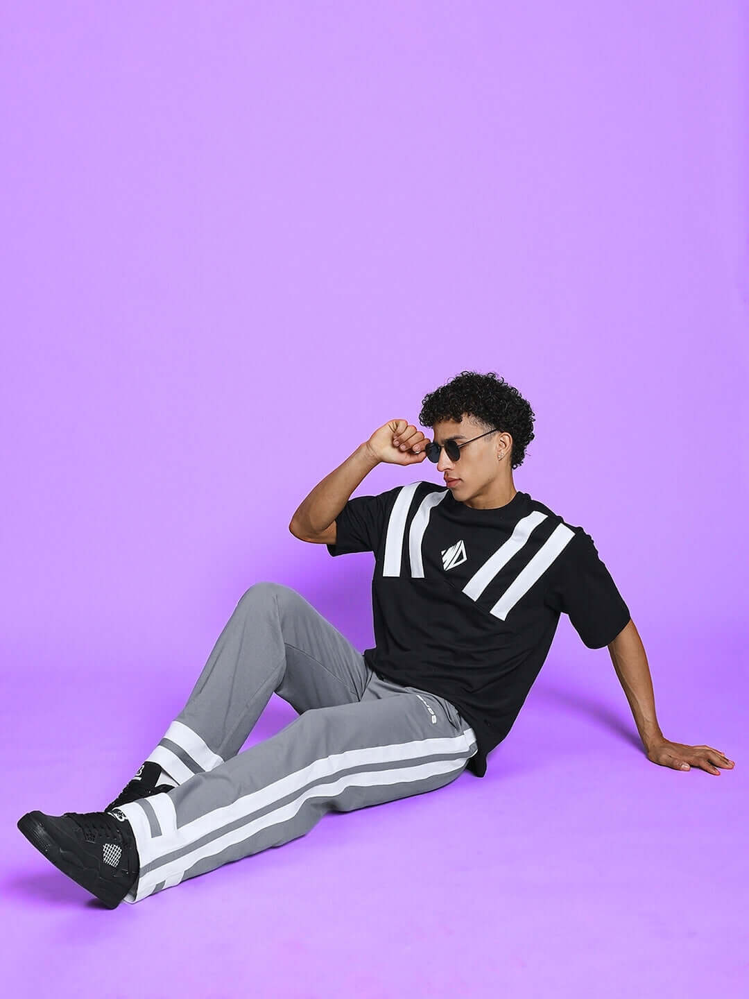 Model wearing grey-white Ronny side stripe jogger, sitting on purple backdrop with black sneakers and sunglasses.