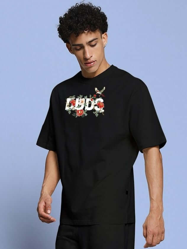 Model wearing ROSE CHARM OVER-SIZED T-SHIRT BLACK with rose graphic on chest, showcasing street style and relaxed fit in premium cotton.