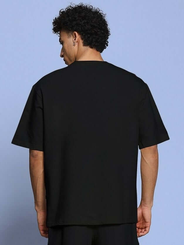 Back view of ROSE CHARM OVER-SIZED T-SHIRT BLACK on a model against a blue background.