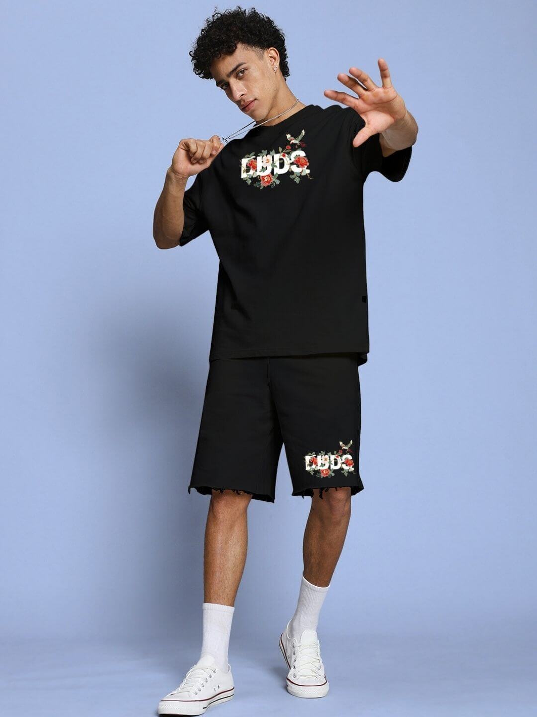 Man wearing ROSE CHARM OVER-SIZED T-SHIRT BLACK with rose graphic, styled in street fashion with matching shorts, on blue background.