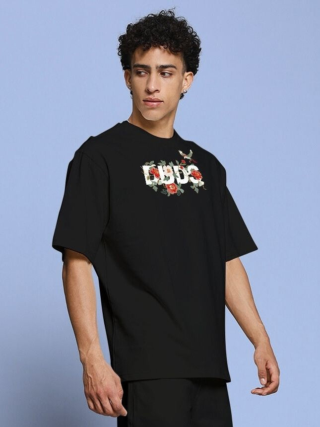 ROSE CHARM OVER-SIZED T-SHIRT BLACK worn by male model, featuring floral logo on chest, casual streetwear fashion.