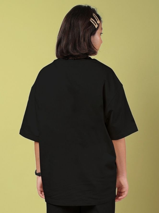 Back view of ROSE CHARM OVERSIZED T-SHIRT FOR BOYS AND GIRLS BLACK worn by a young person against a yellow background.