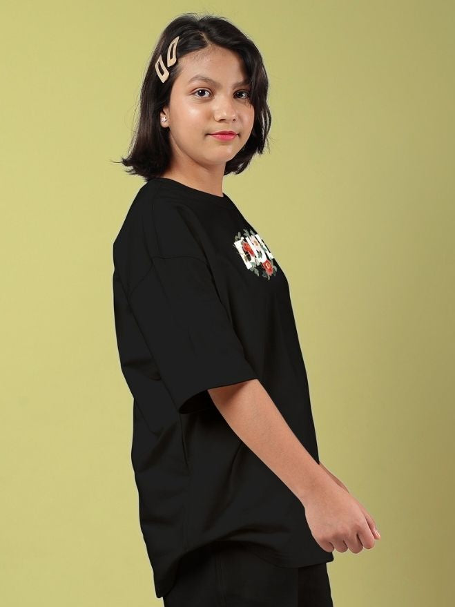 ROSE CHARM OVERSIZED T-SHIRT FOR BOYS AND GIRLS BLACK worn by a young person standing against a yellow background.