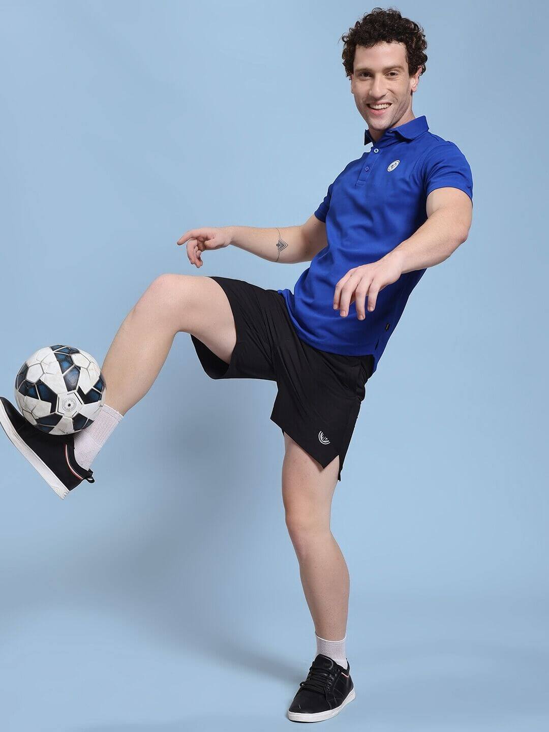 Man playing soccer wearing ROSEWOOD RELAXED FIT SHORTS BLACK and blue polo on light blue backdrop.