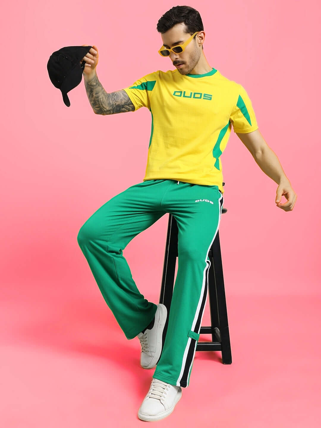Man wears ROSSTON FLAT-OUT RELAXED FIT JOGGERS GREEN with sporty yellow shirt, seated against pink background, showcasing casual street style.
