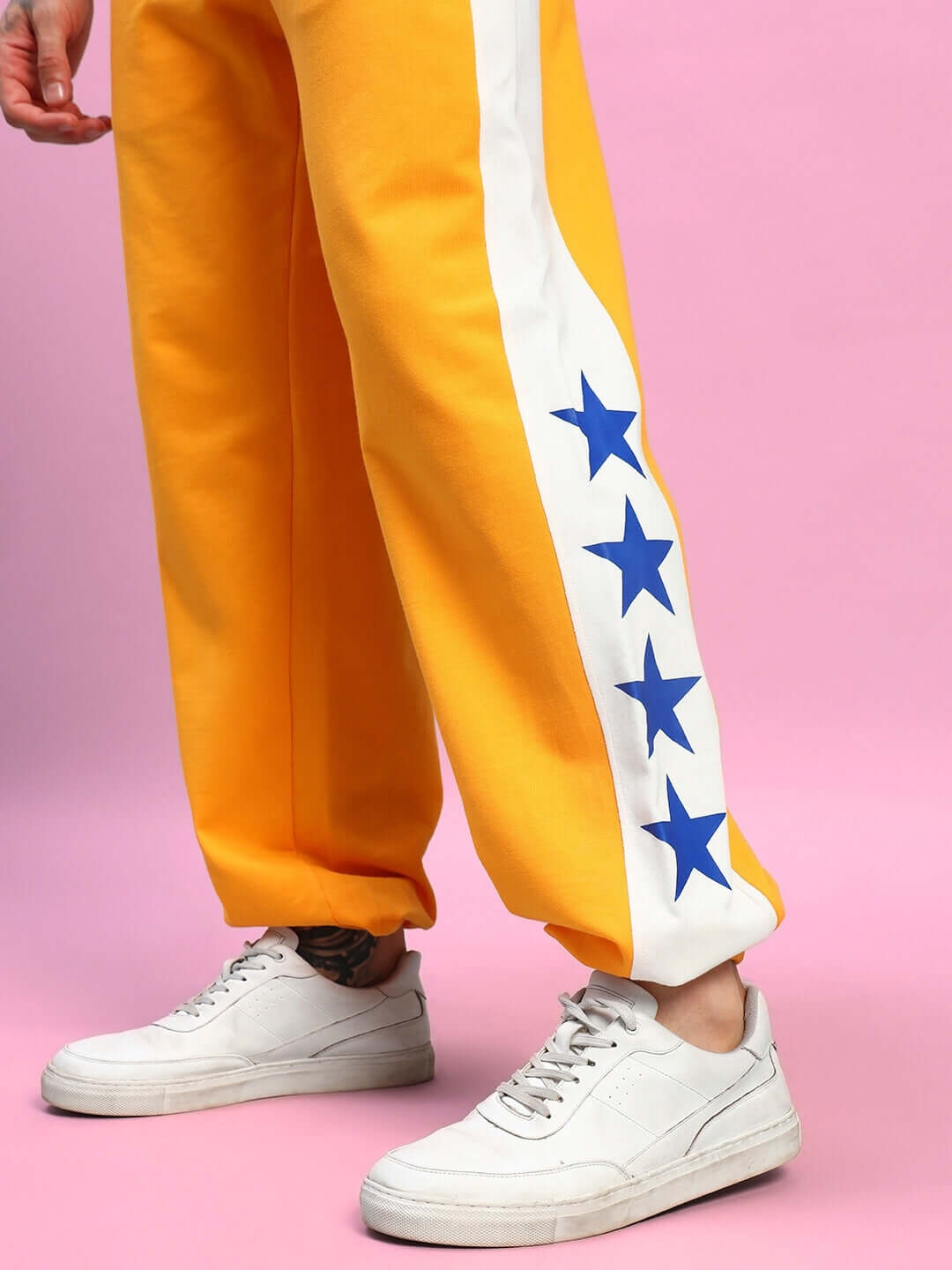 Orange joggers with white side stripe and blue star design, relaxed fit. Modeled with white sneakers against a pink background.