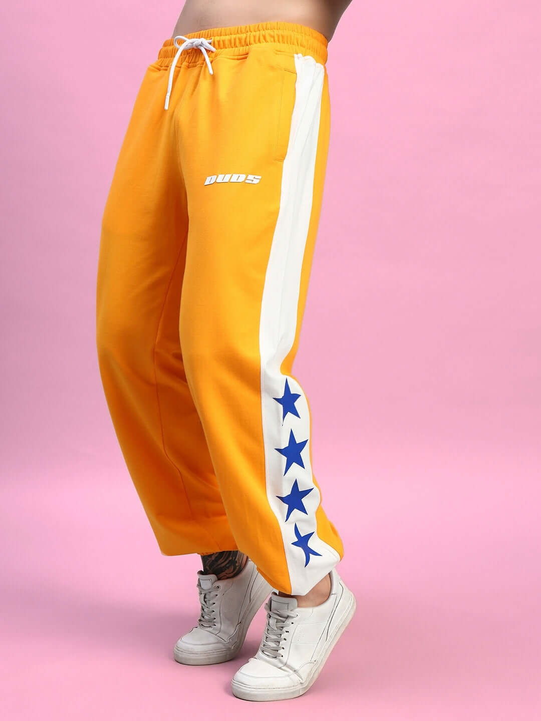 ROX Primer Relaxed Fit Joggers in orange with white star-accented stripe, worn with white sneakers on a pink background.