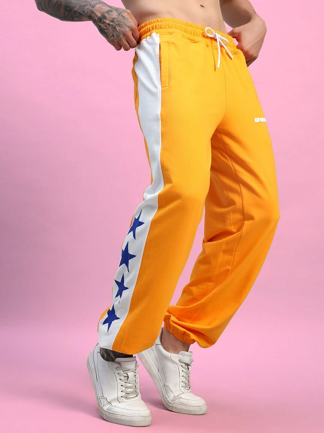 Orange and white relaxed fit joggers with star design detail, worn by a model standing on a pink background.