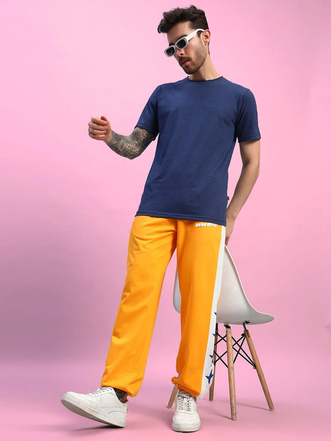 Model wearing ROX Primer Relaxed Fit Joggers in orange-white, paired with a navy shirt and white sneakers, against a pink background.