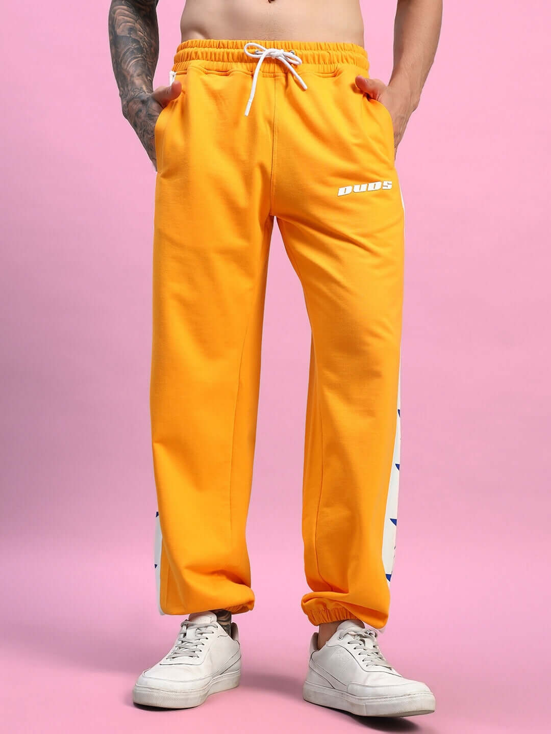 Model wearing ROX PRIMER Relaxed Fit Joggers in Orange-White with white sneakers against pink background.