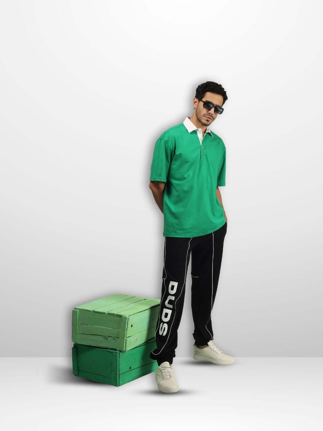 Model wearing SAGOL green oversized polo shirt with black pants, standing beside green boxes. Casual, comfortable look for everyday wear.