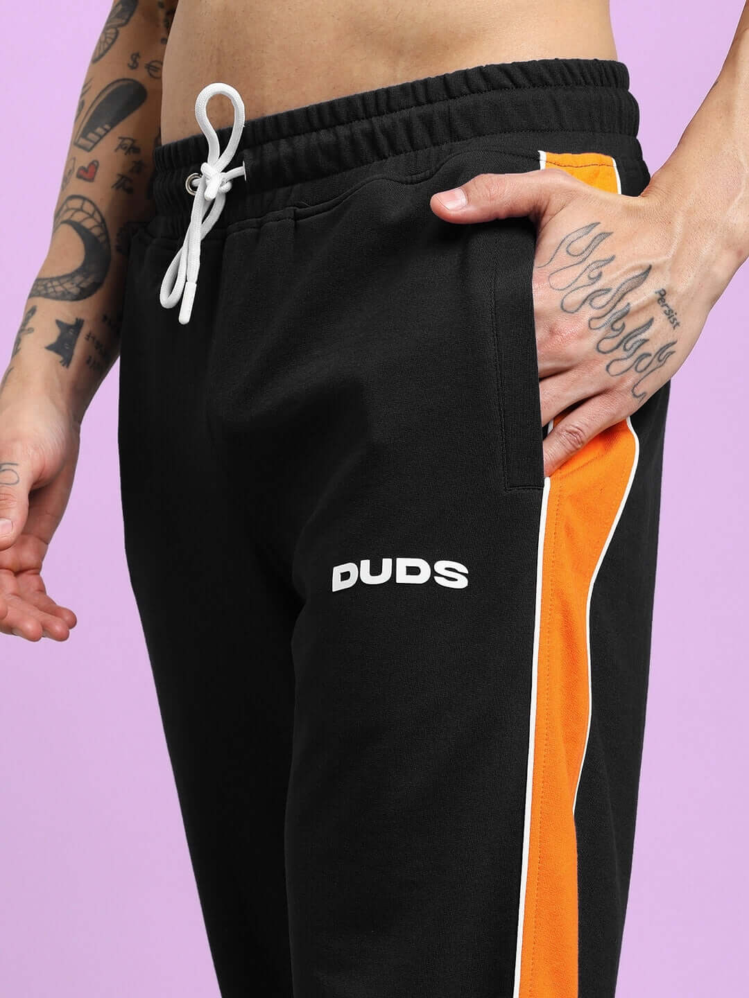 Close-up of SANFORD STRIPE JOGGERS IN BLACK with bold orange side stripes and white drawstring, worn by a tattooed model's hand in pocket