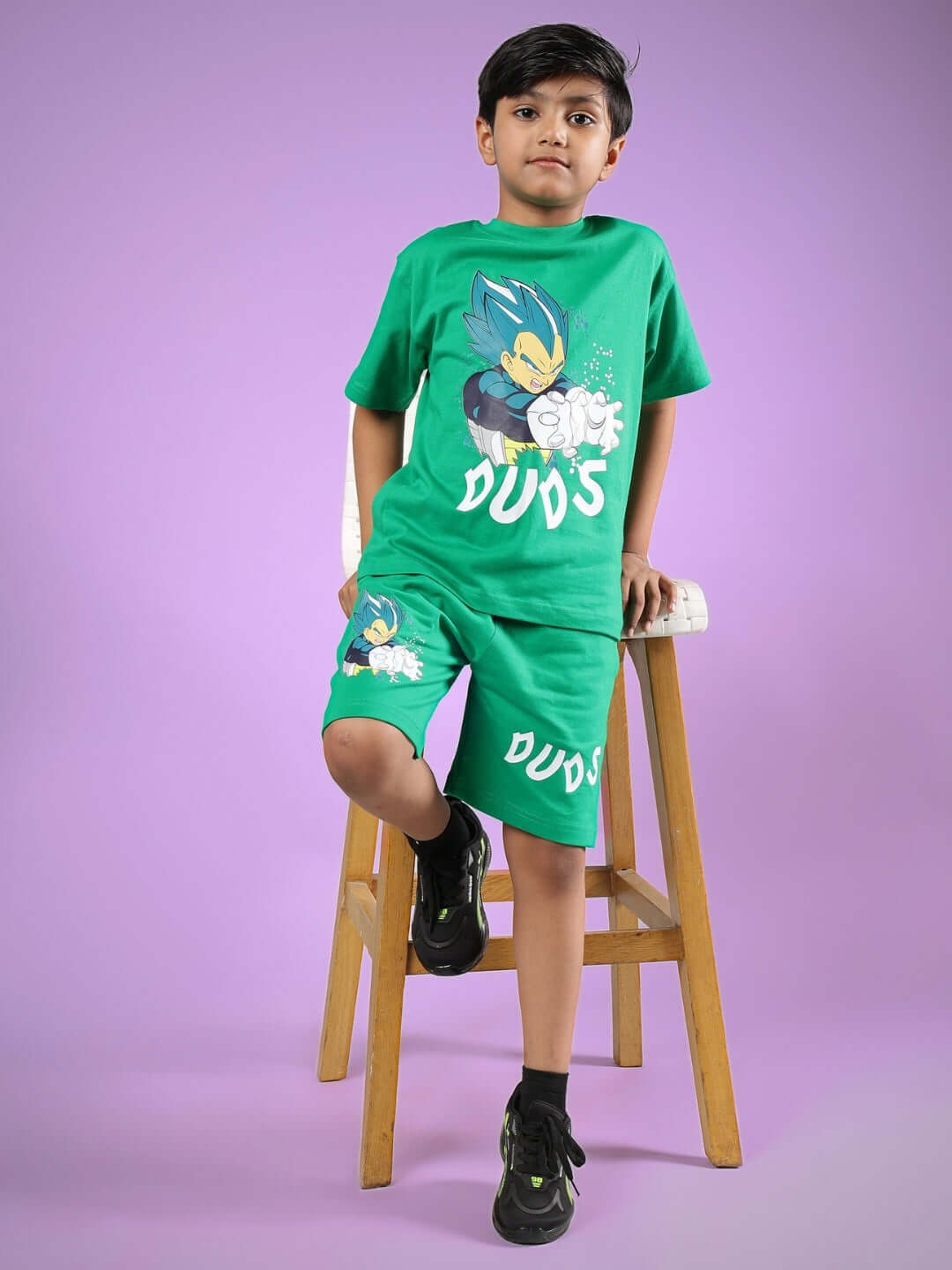 Boy wearing green SATORU SHORTS FOR BOYS & GIRLS GREEN outfit, sitting on a stool in a playful pose.