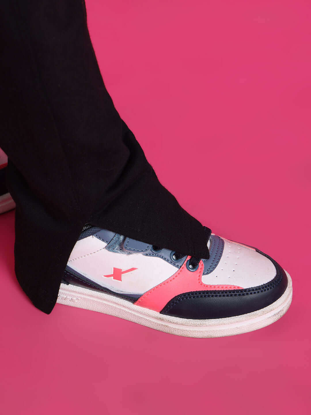 Close-up of a sneaker in pink and navy accents, styled with black scooter co-ord pants for boys and girls on a pink background.