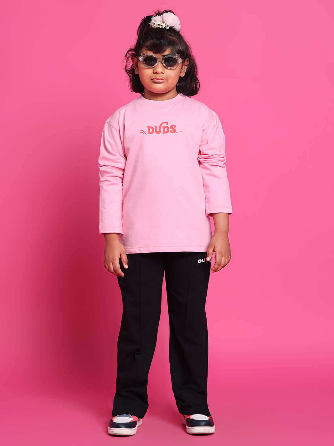 Child wearing DUDS pink scooter co-ord for boys & girls, long-sleeve top and black pants, stylish sunglasses, on a pink background.