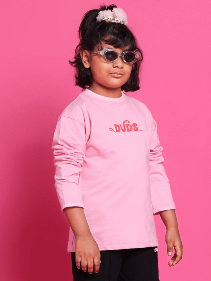 Child wearing pink oversized sweatshirt from DUDS, featuring long sleeves and stylish sunglasses, against a pink background.